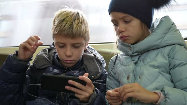 Children with headphones are watching videos on mobile phone on city underground train. Internet technology for streaming video on mobile phone in underground transport. Conscious use free time on way