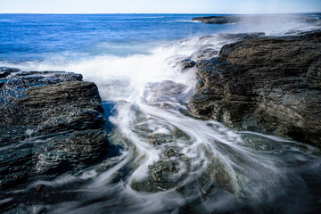 Smooth silky waves flow over the black rocks on the beach. Panoramic motion blur rocky shore image.