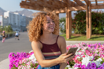 young redhead with curly hair and jeans sitting in a square taking a selfie smiling among pink and violet flowers.