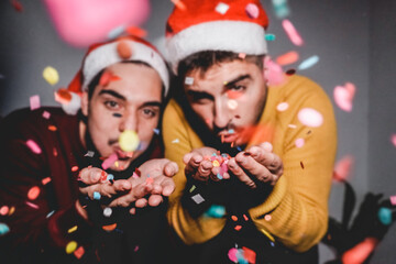 Gay couple celebrating Christmas blowing confetti wearing Santa Claus hats - Flash on 35mm vintage...