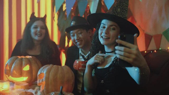 A group of friends are sitting at a table in creepy costumes, taking pictures on their phone and laughing on Halloween