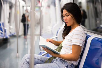 Asian woman sitting on bench in subway train and reading notes in her notebook.