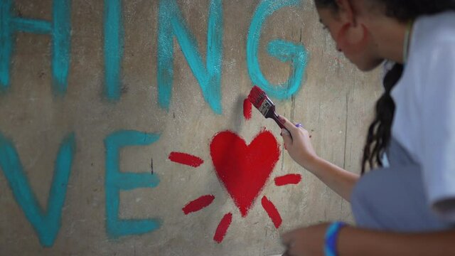 Close up cinematic shot of a white caucasian girl painting a red hearth and a beautiful quote on the wall of an African school. Love concept. Do everything with love. Tanzania, Africa 4K.