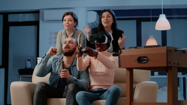 African american woman using vr glasses and controllers to play video games with workmates after work. Colleagues enjoying free time with alcoholic drinks after hours, playing game on tv