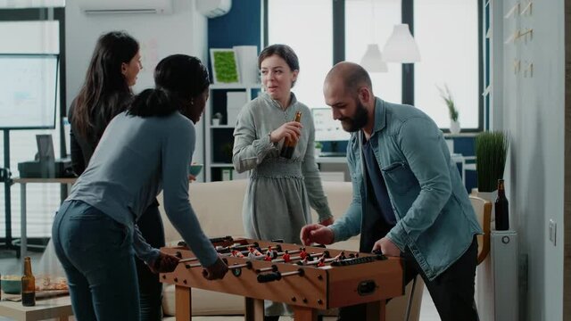 Diverse group of workmates enjoying game with foosball table to do fun activity after work. Coworkers playing football and drinking beer from bottles, celebrating party with entertainment