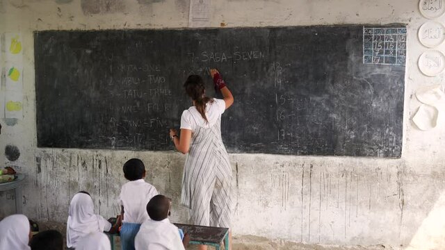 White caucasian teacher writing on the blackboard numbers teaching black African children. NGO cooperation volunteering helping for the development of  education in Africa 4K.