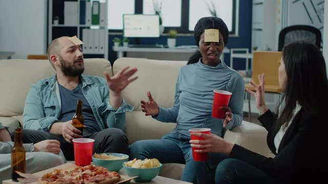 Happy colleagues playing guessing game for fun activity after work. Cheerful workmates enjoying charades play with sticky notes on forehead for entertainment with drinks after hours