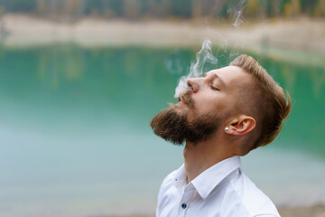 Business caucasian young bearded man in a white shirt smokes a cigarette and blows smoke against...