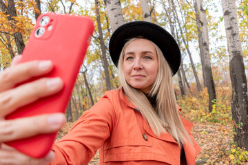 Young girl with blond hair makes a selfie on the phone while standing in the autumn forest.