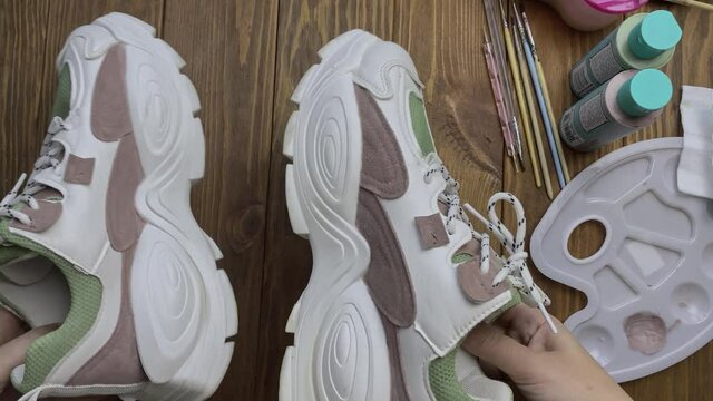 Person's hands take colored sneakers and twist it in front of camera. Plastic palette, acrylic paints, brushes and napkins on wooden table. Shoe design, hobbies and leisure at home.