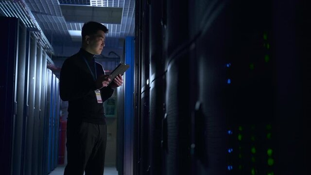 IT specialist using device and doing network diagnostics while standing on render farm spbas. Young Asian man holds device in hand and inspects equipment racks or hardware, thinks about something and