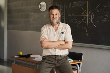 Middle-aged bearded male teacher looking at camera with arms crossed in classroom.