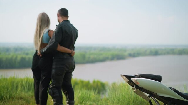 Man driver with beloved girlfriend embraces near modern motorcycle standing on hill top grass against calm lake on spring day backside view slow motion