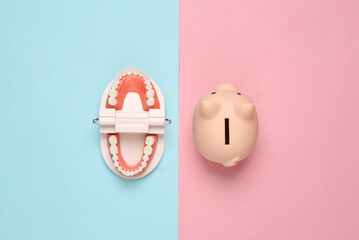 Model of human jaw with piggy bank on pink blue pastel background. Creative flat lay