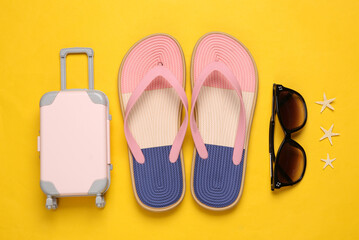 Colored flip flops with luggage and sunglasses on yellow background. Beach vacation concept. Flat lay
