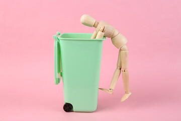 Wooden puppet rummages in a trash can on a pink background. Poverty concept