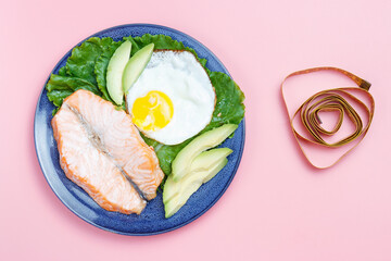 Plate with fish and vegetables, tape measure on pink background.