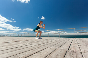 Fit woman doing squat exercise with fitness elastic bands, training legs on the embankment at bright sunny day with blue sky and clouds