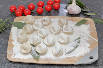 handmade dumplings on a wooden board are sprinkled with flour, next to tomatoes, bay leaf, garlic