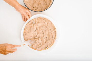 Culinary confectionery hobby. Top view female cook's wrinkled hands decorating cake with cream filling using spatula, housewife preparing delicious layered cake on white table indoors, copy space