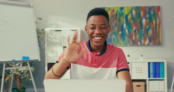 A young boy of African American beauty sits at desk in an office room with a laptop, smiles broadly and waves hand, carries on a conversation via internet, video, has remote teaching