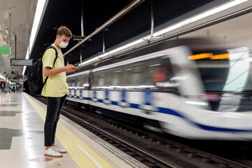 Young man with mask looking at his cell phone in a subway station as a carriage speeds by
