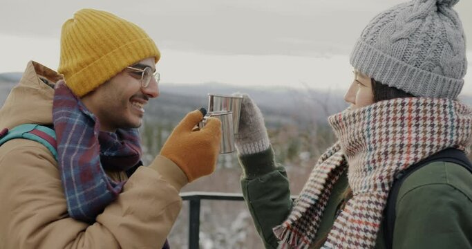 Happy couple tourists clinking metal cups drinking laughing and watching amazing winter landscape mountains and forest. People and tourism concept.