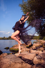 Concepts of Dance. Young Brunette Female Ballerina In Black Dress Showing Dance Pas on Rocky Shore While Wearing Seashell Decorated Crown And Pearl Necklace.