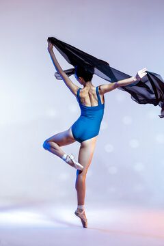 Classical Ballet Ideas. Young Japanese Female Ballet Dancer Dancing With Flying Black Cloth While Posing in Blue Bodysuit Against White Background.