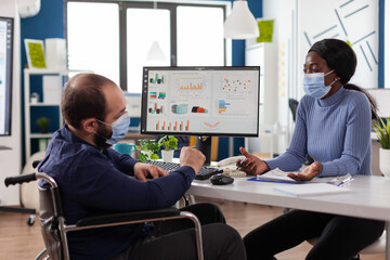 Fototapeta na wymiar Paralyzed businessman in wheelchair wearing protective face mask to prevent infection with coronavirus working in startup business office. Multi-ethnic workteam discussing company strategy