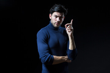 Thinking Handsome Brunet Man Posing in Blue Turtle Neck Sweater With Lifted Hand Against Black Background With Copy Space as Successful Young Man.
