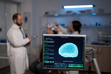 Monitor with brain activity on screen in neurology medical laboratory. In background team of neurologist adjusting eeg headset of woman patient monitoring nervous system during neuroscience experiment - obrazy, fototapety, plakaty