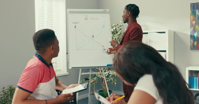An experienced long-term employee of large corporation stands in office by white board draws chart conducts training for trainees, young future employees take notes while listening to dark-skinned men