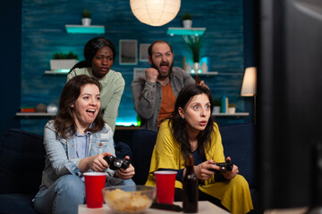 Excited women friends sitting on sofa winning videogames using gaming joystick playing games during online competition. Multi-ethnic group of people enjoying spending time together. Friendship concept