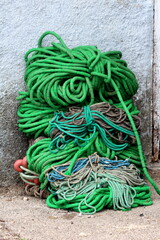 Large pile of multicolor heavily used thick old naval ropes mixed with small red buoys left next to dilapidated house wall on gravel road on warm sunny spring day