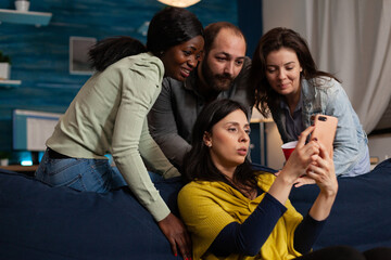 Multi-ethnic friends looking at fuuny videos on smartphone during movie night at home enjoying...