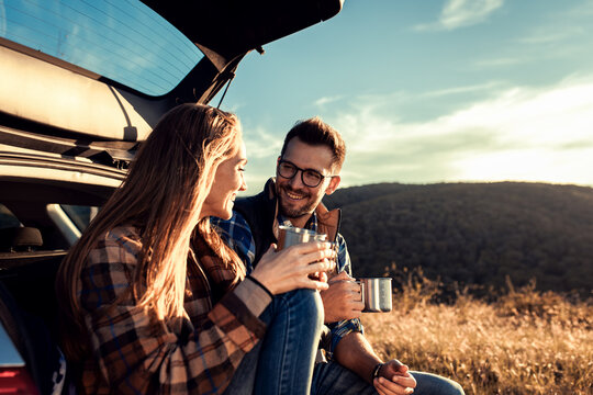 Couple on road trip sitting in trunk of a car resting and drinking coffee.