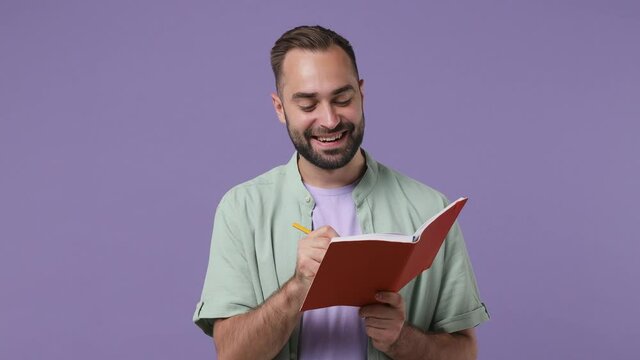 Smiling dreamful pensive thoughtful wistful minded young bearded man 20s years old wears mint shirt hold write fill in brown notebook diary isolated on plain light purple background studio portrait
