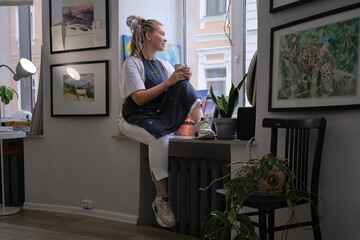 Artist Woman in dirty apron resting with a cup of tea sitting on the windowsill and looks out the window