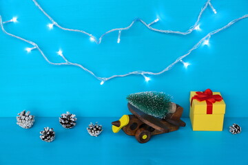 The wooden model of retro airplane with fir tree on is among winter pinecones and gift box on the blue wooden background with garland lights. Concept of Christmas, New Year, holiday, vacation, travel.