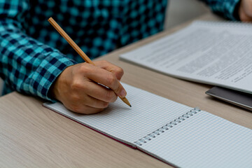 A woman in a plaid shirt writes with a pencil in a notebook and looks into a paper report. The right female hand is in focus. Horizontal view.