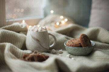 A cup of hot coffee with marshmallows and chocolate chip cookies on the blanket with christmas lights