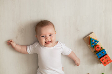 Fototapeta premium cheerful newborn baby in a white bodysuit lies on his back on the floor and plays with educational toys. products for children. concept of a happy childhood and motherhood. child care. space for text
