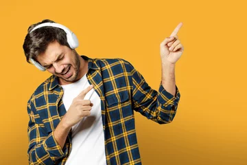 Tuinposter Portrait of handsome beard guy in white t-shirt and plaid shirt listening to music in headphones singing and pointing fingers aside isolated on yellow orange background © Paulista