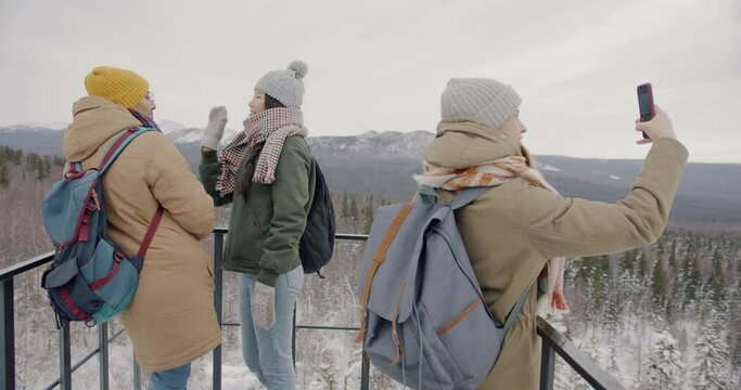Happy tourists watching amazing landscape of winter mountains from observation deck and taking pictures with smartphone. People and modern technology concept.