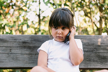 Little girl with black hair, wearing a white t-shirt, sitting on a bench in a park talking on the mobile phone, with a serious gesture.