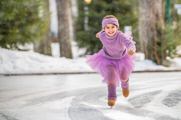 little girl in pink sweater and full skirt rides on sunny winter day on an outdoor ice rink in park