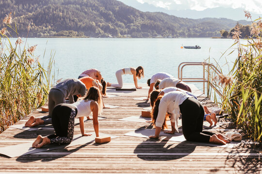 Female instructor teaching yoga to men and women on jetty by lake