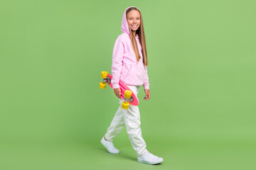Fototapeta na wymiar Full size photo of nice small blond girl hold skate wear hoodie trousers sneakers isolated on green background