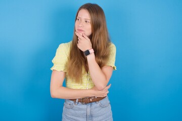 Shot of contemplative thoughtful young ukranian girl wearing yellow t-shirt over blue backaground  keeps hand under chin, looks thoughtfully upwards.